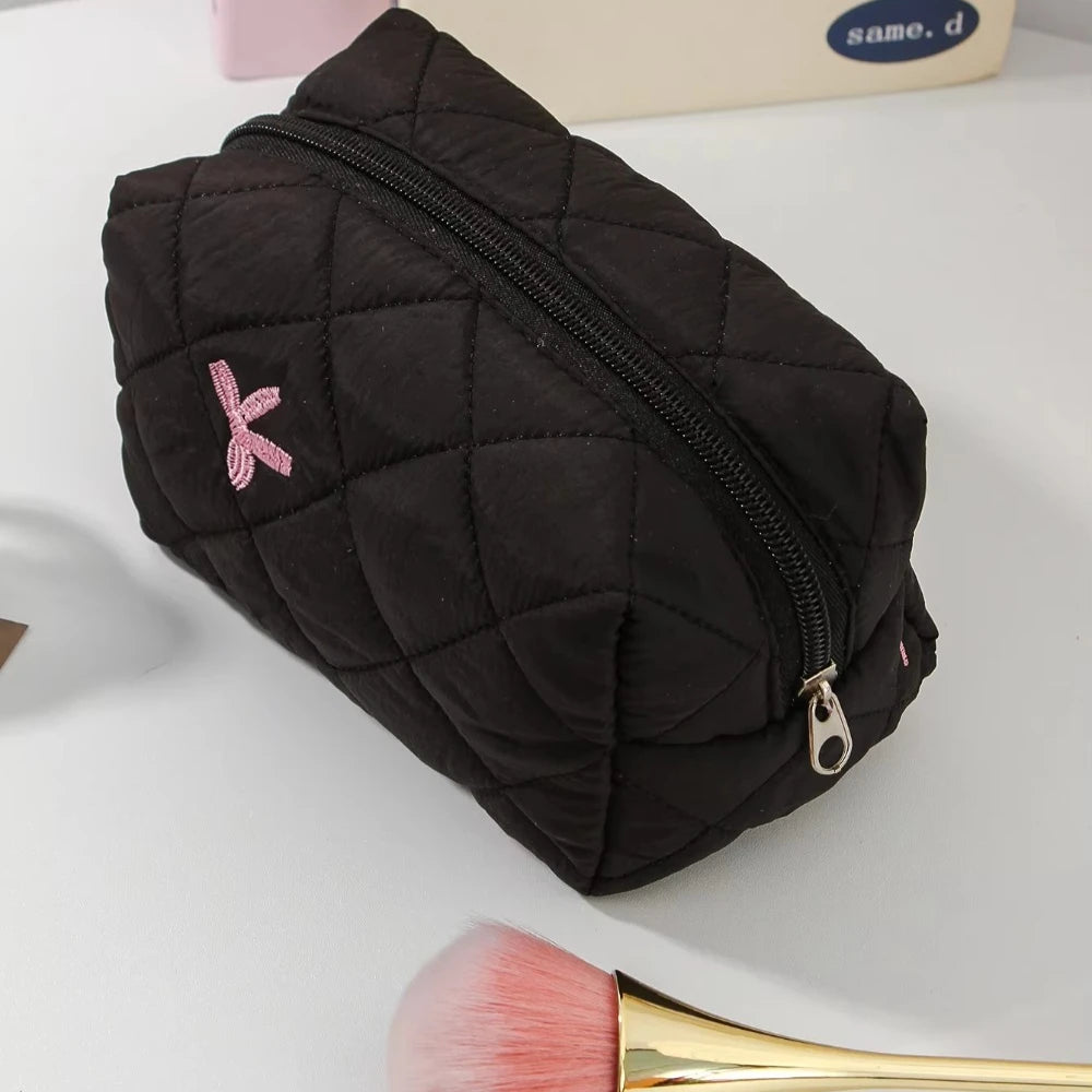 A black quilted cosmetic bag with a zipper and a pink bow detail is shown next to a makeup brush.