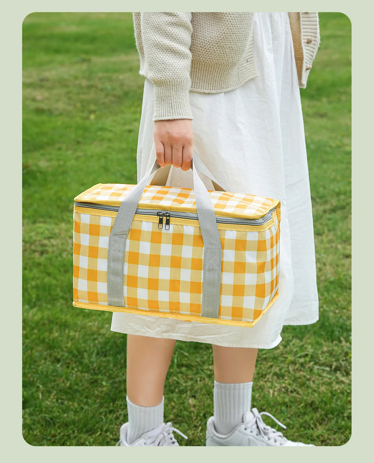 A person holds a yellow and white checkered insulated picnic tote bag outdoors.