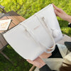 Bolso tote de cuero PU blanco con dije plateado y pompom esponjoso, sostenido por una persona al aire libre.