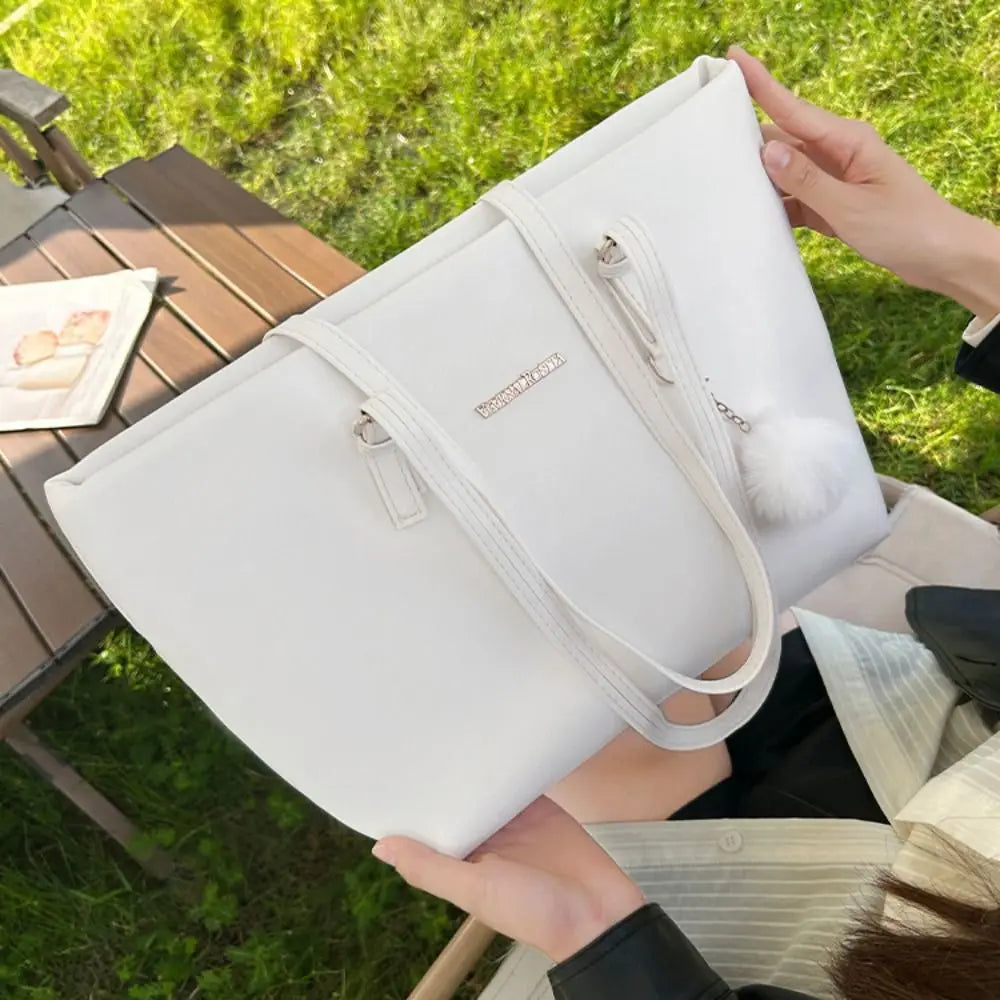 Bolso tote de cuero PU blanco con dije plateado y pompom esponjoso, sostenido por una persona al aire libre.