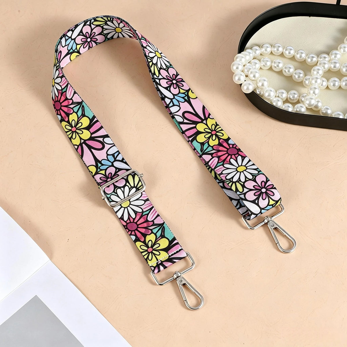 A floral patterned nylon crossbody bag strap with metal buckles is displayed next to a pearl necklace.