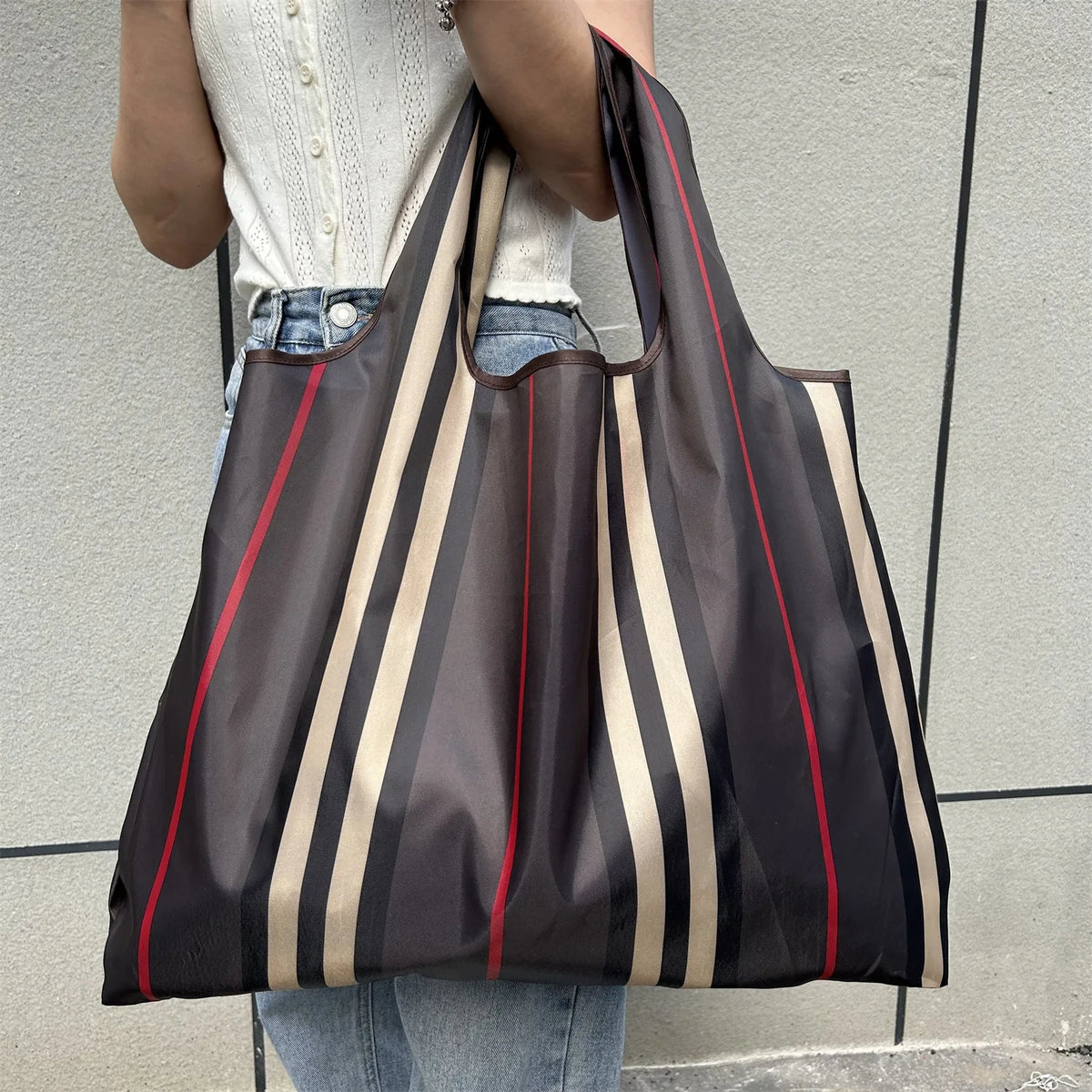 A person holds a dark brown and beige striped foldable oxford grocery tote bag against a gray wall.