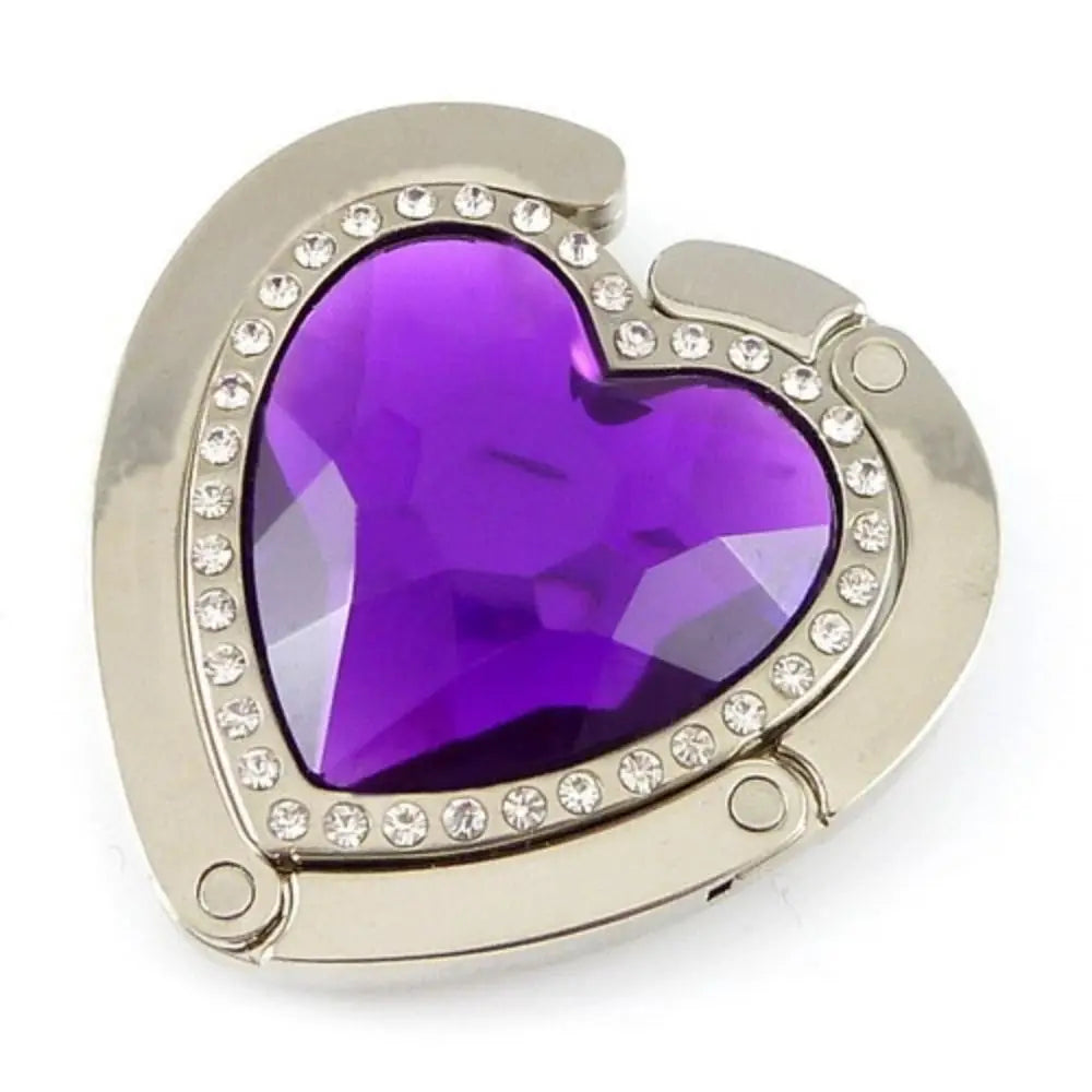 A heart-shaped metal fold-over bag hook with a purple gemstone set in a silver-toned frame.