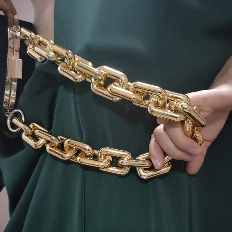 Una mano sostiene una cadena de bolso acrílico de color dorado contra una tela verde oscuro.