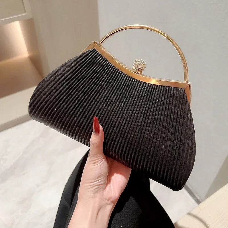 A black pleated evening handbag with a gold top handle is held by a hand against a light background.