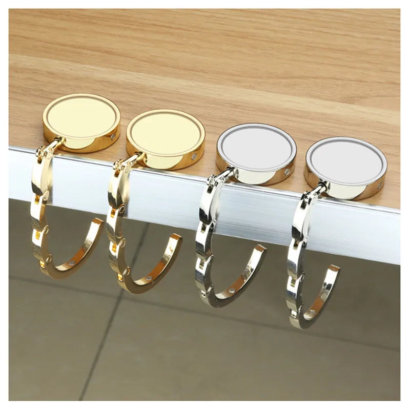 Four gold and silver folding alloy handbag hangers displayed on a wooden surface.