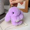 A fluffy purple plush rabbit charm is shown being held by a hand, resting on a piece of paper.
