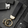 A close-up of a gold-toned carbon fiber leather keychain attached to a black surface.
