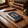 A vintage metal card holder with an engraved gold design rests on a leather-bound book and wooden surface.
