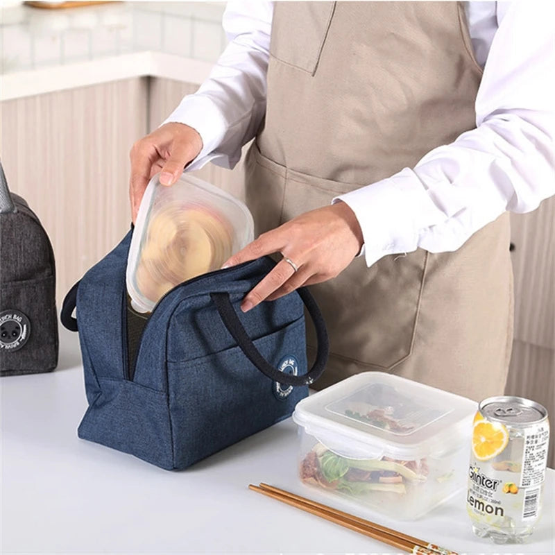 A person puts a food container into a dark blue insulated lunch bag while wearing a beige apron.
