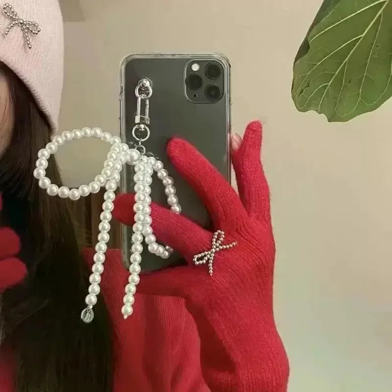 A hand wearing a red glove holds a pearl necklace with a bow charm and a heart pendant against a light background.