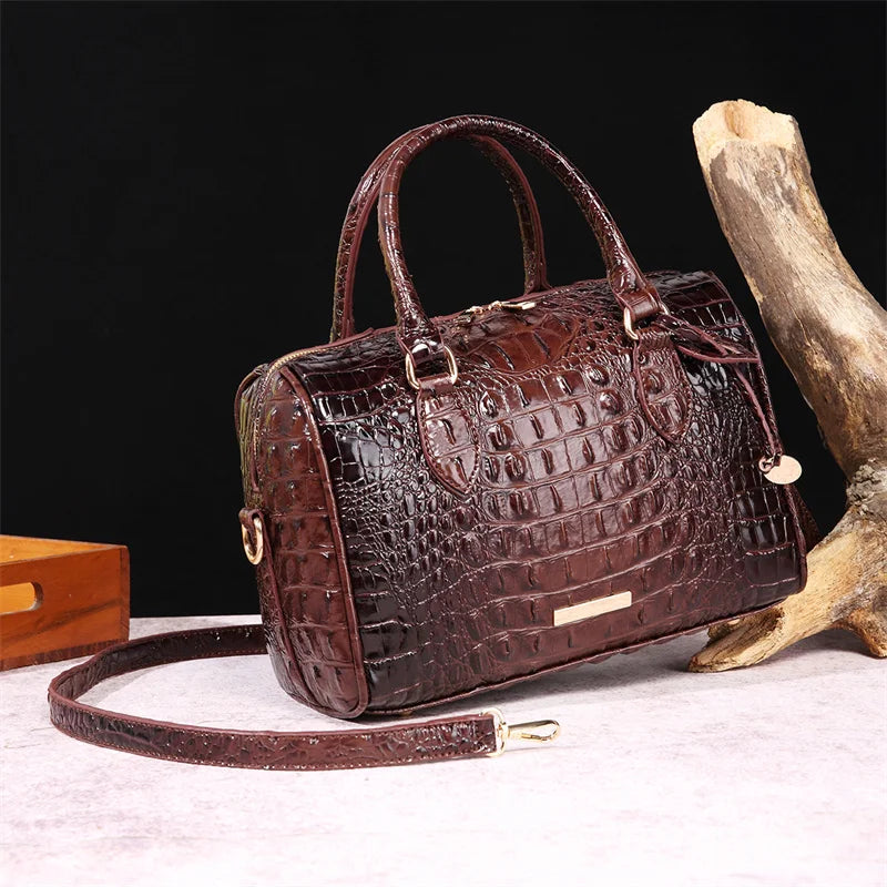 A dark brown crocodile pattern leather crossbody tote bag resting on a light surface next to a piece of driftwood.