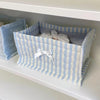 A light blue and white striped quilted cotton storage basket with a white bow on a white shelf.