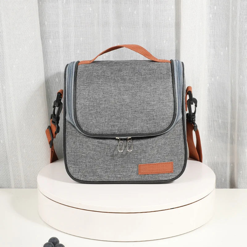 A gray insulated reusable lunch bag with brown straps resting on a white circular stand.