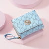 A light blue elegant letter zipper wristlet wallet with a gold monogram detail resting on a pink surface.