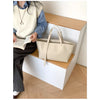 Un sac à main en cuir PU beige et décontracté est exposé sur une étagère en bois à côté d'une personne lisant un livre.