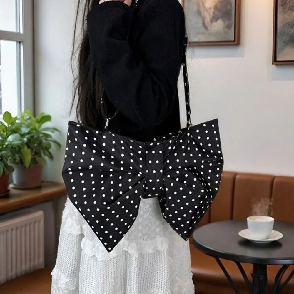 A person is holding an elegant black and white polka dot pleated bow crossbody tote bag against a light background.