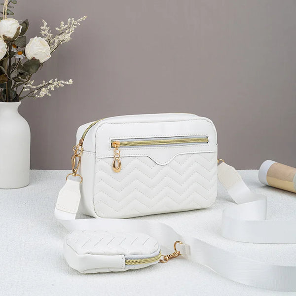 A white quilted pattern crossbody bag with a gold zipper and strap is displayed on a white surface.