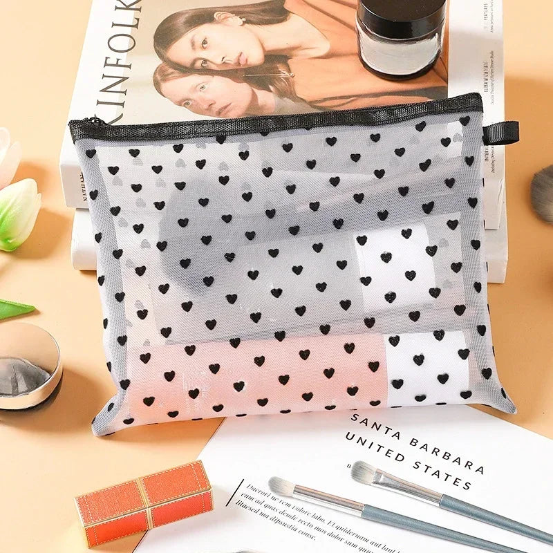 A clear mesh cosmetic storage pouch with a pattern of black hearts and pink dots is shown resting on a book.