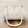 A white satin clutch bag with a pearl chain strap and a pearl-encrusted clasp resting on a wooden surface.