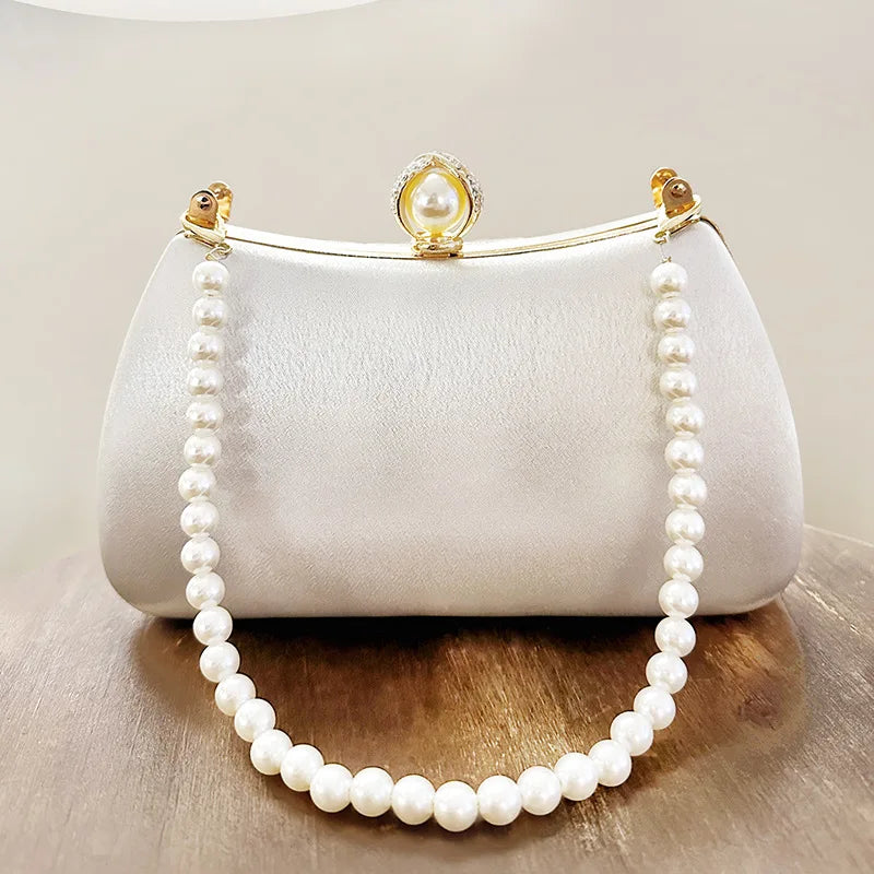 A white satin clutch bag with a pearl chain strap and a pearl-encrusted clasp resting on a wooden surface.