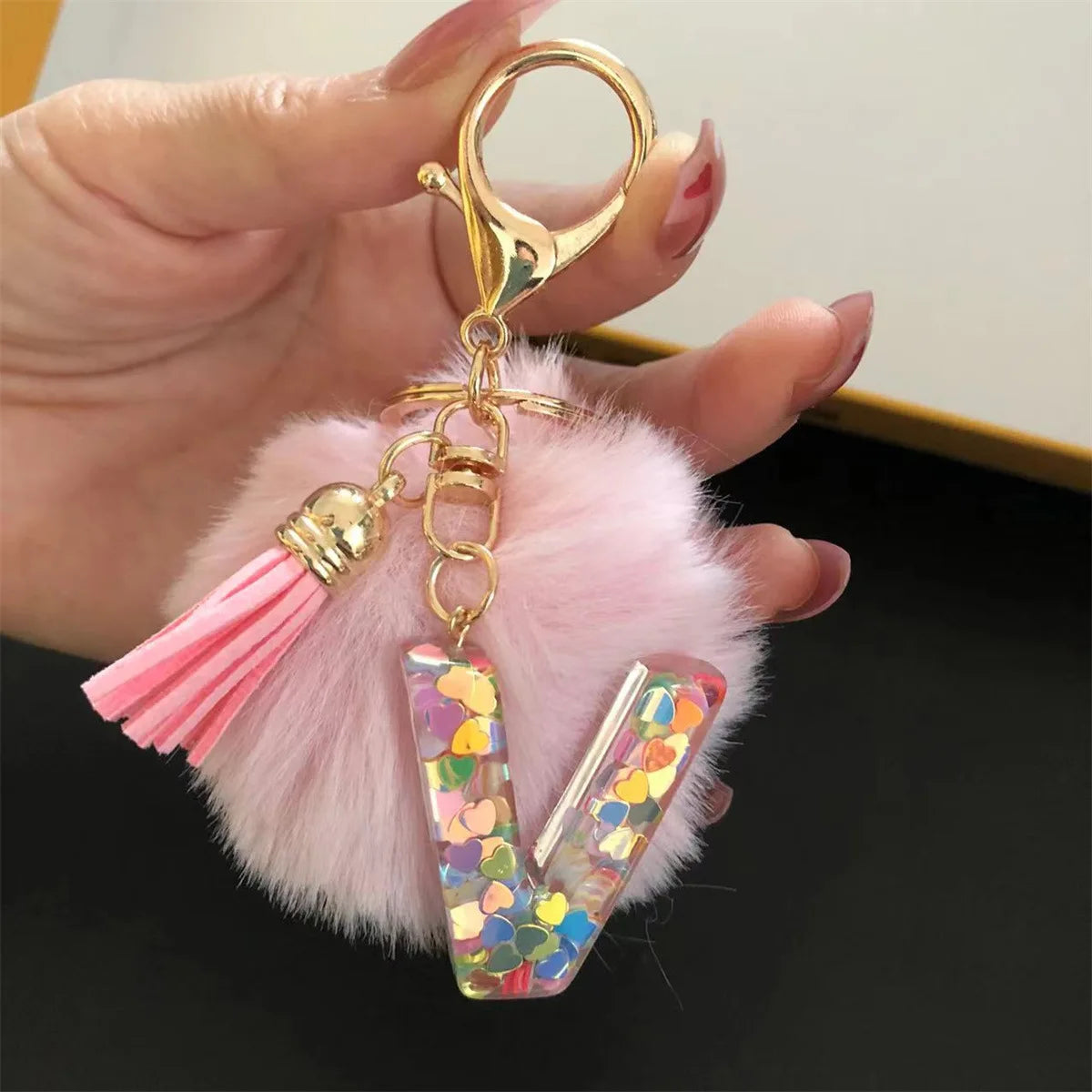 A hand holds a gold initial letter keychain with a shiny resin pendant and pink alphabet charm resting on fluffy pink faux fu