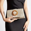 A woman holds a woven straw envelope clutch with an orange beaded strap against a black dress.