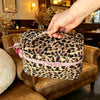 Una mano sostiene una bolsa de almacenamiento con estampado de leopardo y detalles rosas sobre un sofá de cuero marrón.