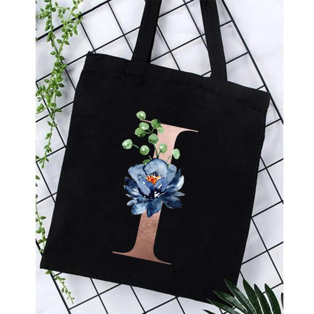Black canvas tote bag featuring a large gold letter I with a blue floral print against a white background.