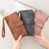 Three leather trifold clutch wallets in brown, gray, and pink colors being held by hands.