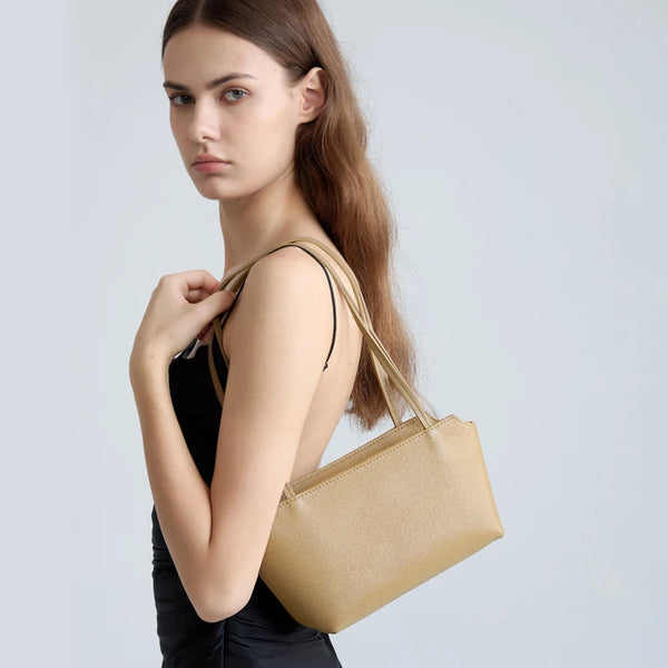 A model holds a gold leather mini shoulder bag with adjustable straps against a light gray background.