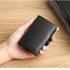 A hand holds a black carbon fiber patterned ID card holder wallet on a wooden surface.