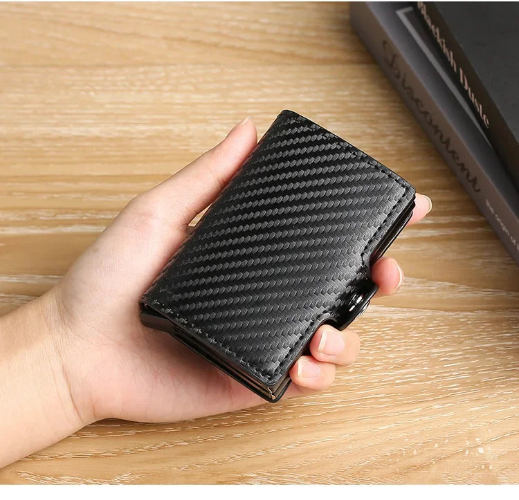 A hand holds a black carbon fiber patterned ID card holder wallet on a wooden surface.
