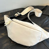 A cream-colored nylon crossbody sling bag is displayed on a dark leather surface.