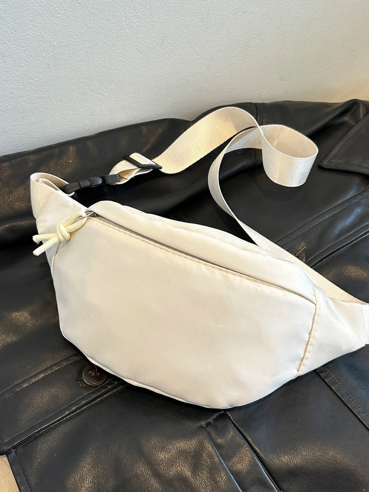 A cream-colored nylon crossbody sling bag is displayed on a dark leather surface.