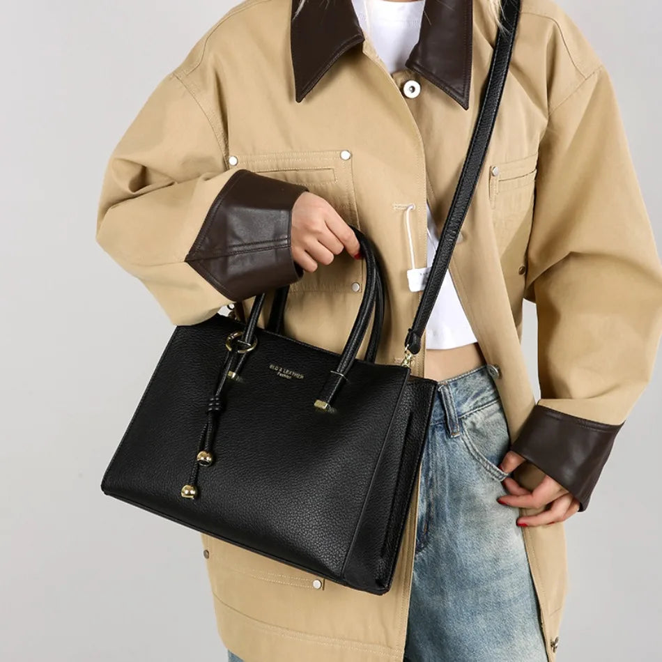 Eine Frau hält eine schwarze Leder-Tasche mit goldener Hardware unter einem beigen Jacken.