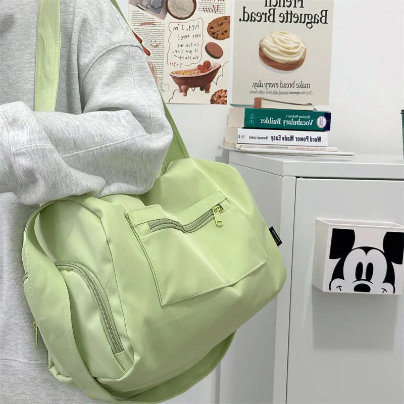 A light green canvas crossbody travel tote bag is shown next to a gray hoodie and some printed materials.