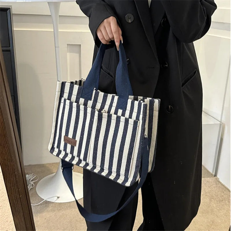 Une personne tient un sac bandoulière en toile rayée bleu marine et blanc sur un fond clair.
