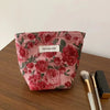 A pink floral cosmetic pouch with a small white label rests on a brown wooden surface next to makeup brushes.