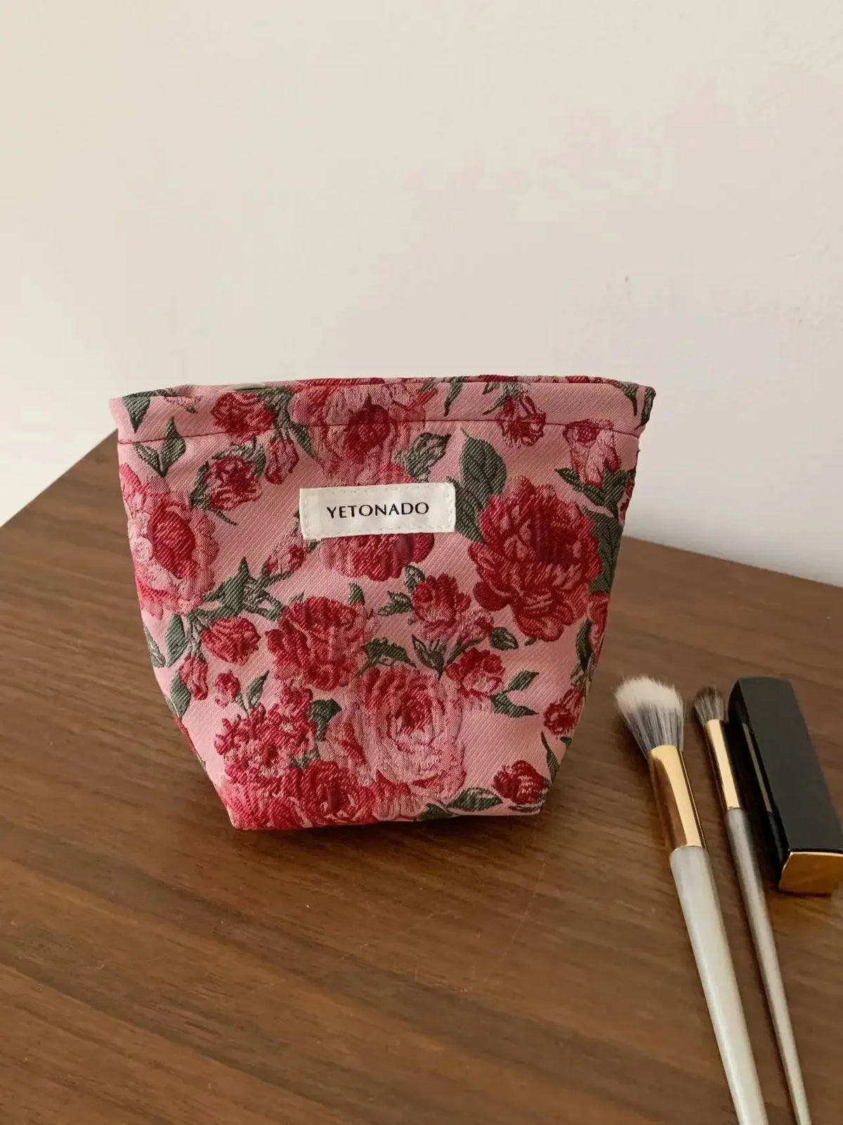 A pink floral cosmetic pouch with a small white label rests on a brown wooden surface next to makeup brushes.