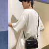 A young man wearing a white shirt and dark hat holds a black leather crossbody bag against a light-colored wall.