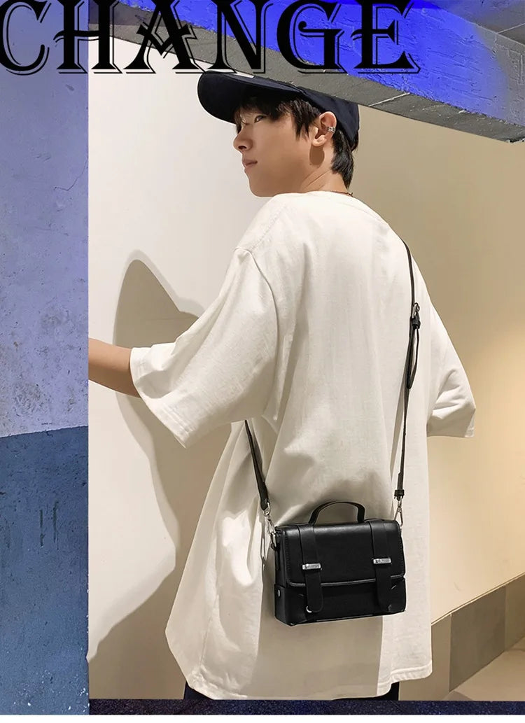 A young man wearing a white shirt and dark hat holds a black leather crossbody bag against a light-colored wall.