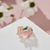 Gold-toned flower butterfly rings with pink and green enamel set against a soft pink background.