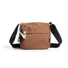 Sac bandoulière à travers en tartan marron et résistant à l'eau avec sangle noire et charm orange.