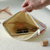 A hand is opening a beige makeup bag with a floral print to reveal wooden makeup brushes inside.