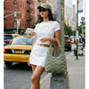 A woman wearing a white t-shirt and skirt stands next to a metal pole holding a green quilted nylon shoulder bag.