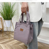 A person holds a mauve insulated lunch bag in a white shirt while standing near a potted plant.
