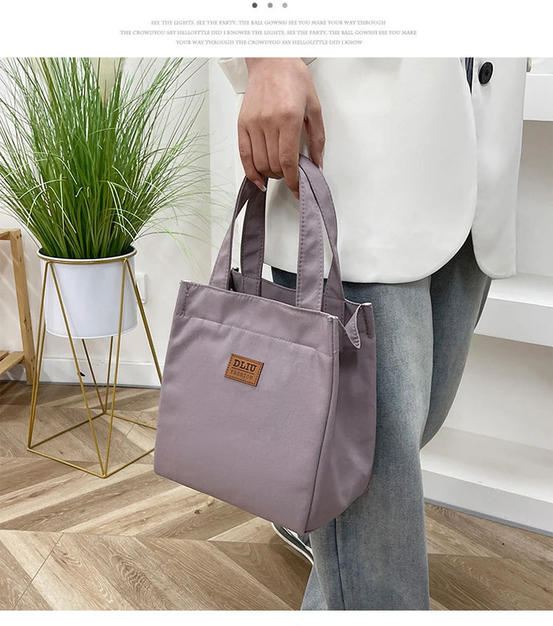 A person holds a mauve insulated lunch bag in a white shirt while standing near a potted plant.