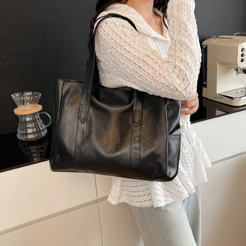 Eine Frau hält eine schwarze Vintage-PU-Leder-Umhängetasche vor einem weißen Schrank mit einer Kaffeemaschine.