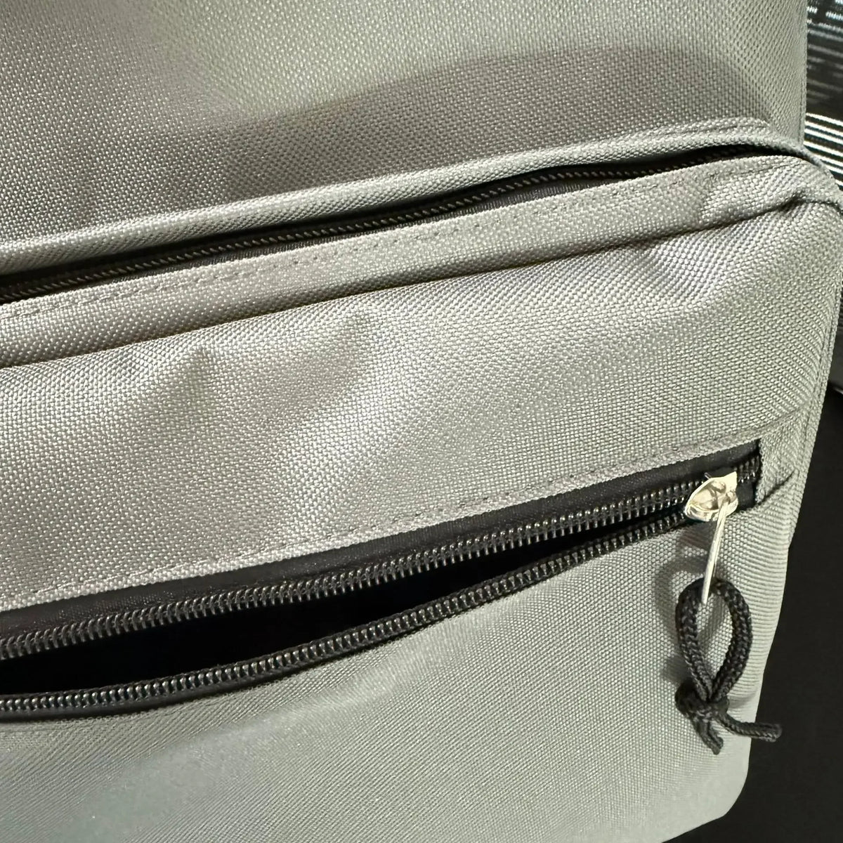 A close-up of a light gray canvas backpack showing a zippered pocket detail.